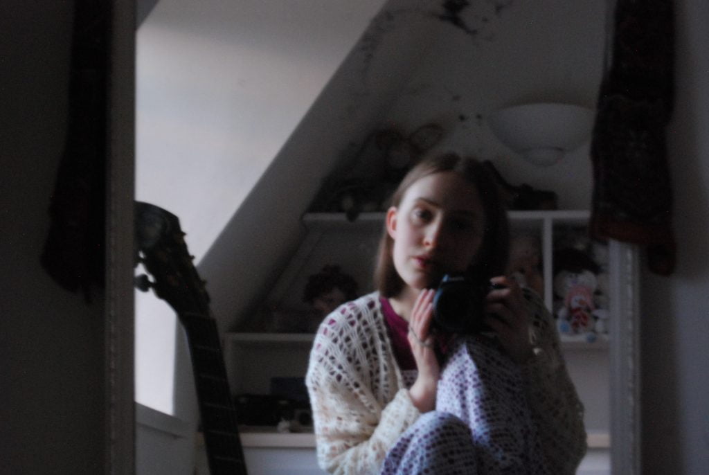 A mirror selfie in which Sakara (a thin white woman with light brown hair) gazes into the camera. The light is grey and fuzzy, she wears a cream crocheted cardigan and purple spotty pyjamas.