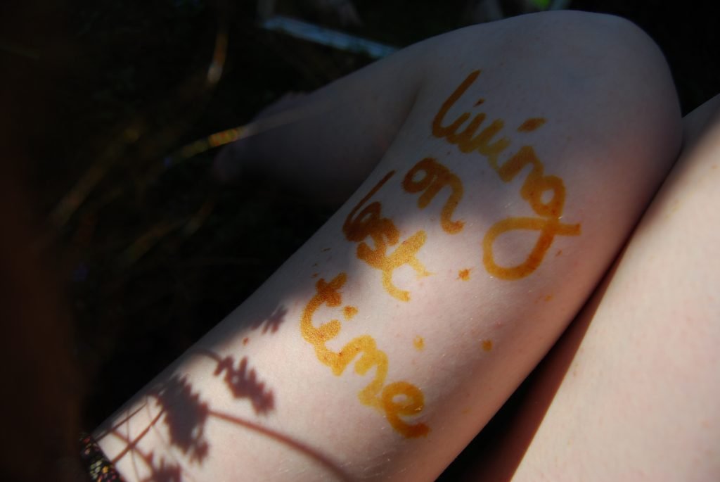 The words "Living On Lost Time" are written in brown on the white skin of Sakara's left thigh. Small flowers cast shadows across the words.