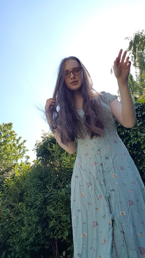 Photo of Sakara wearing a button-front floral dress, she holds back her hair with one hand and partially covers the sun behind her with the other.