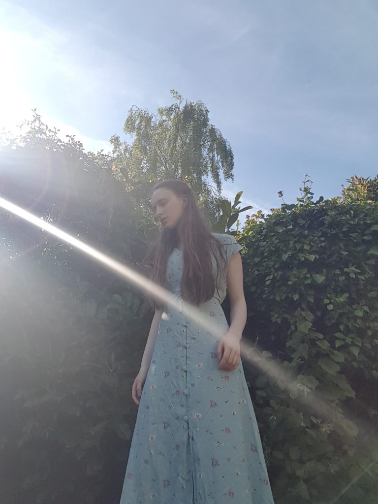 Almost full length photo of Sakara standing in front of green hedges and a blue sky. Wearing the same dress described before.