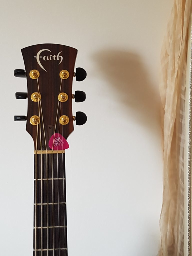 The top (or headstock) of a guitar and the beginning of the neck. There is a pink pick tucked into the strings.