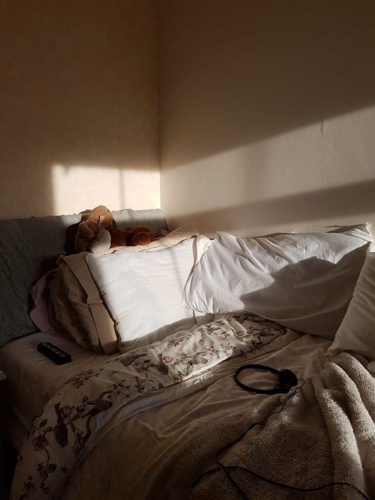 Photograph of a bed in the corner of a room with evening sunlight coming in across parts of the pillows. It feels warm and inviting in beige and cream tones. There are two cuddly toys sitting on top of a pillow (one rabbit and one dog) and a pair of headphones on the bed.