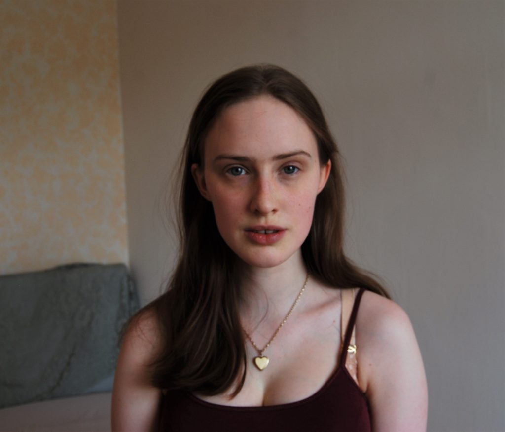 Photograph of Sakara from the chest up, a thin white woman with medium length brown hair looking directly at the camera. There are tired shadows under her eyes and she's not wearing any make up but looks alert. There is a gold heart locket around her neck.