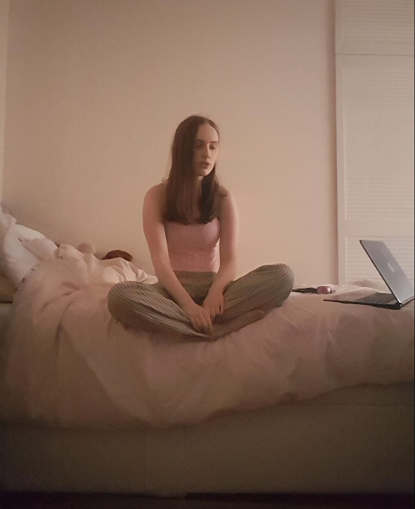 Sakara sits on a bed in a dimly lit room wearing a similar outfit, only with a pink version of the top.