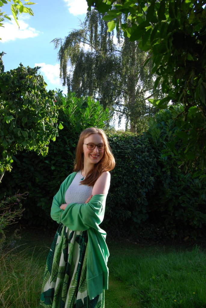Sakara stands with her arms folded, smiling. She's wearing a full 1950s style skirt printed with green leaves, a pale blue top and a bright green cardigan.