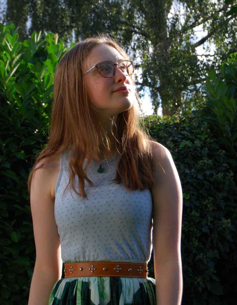 Sakara stands outside in a sunny garden, surrounded by lots of greenery. She has medium length golden brown hair and wears brown mottled glasses. She's smiling slightly and seems content.