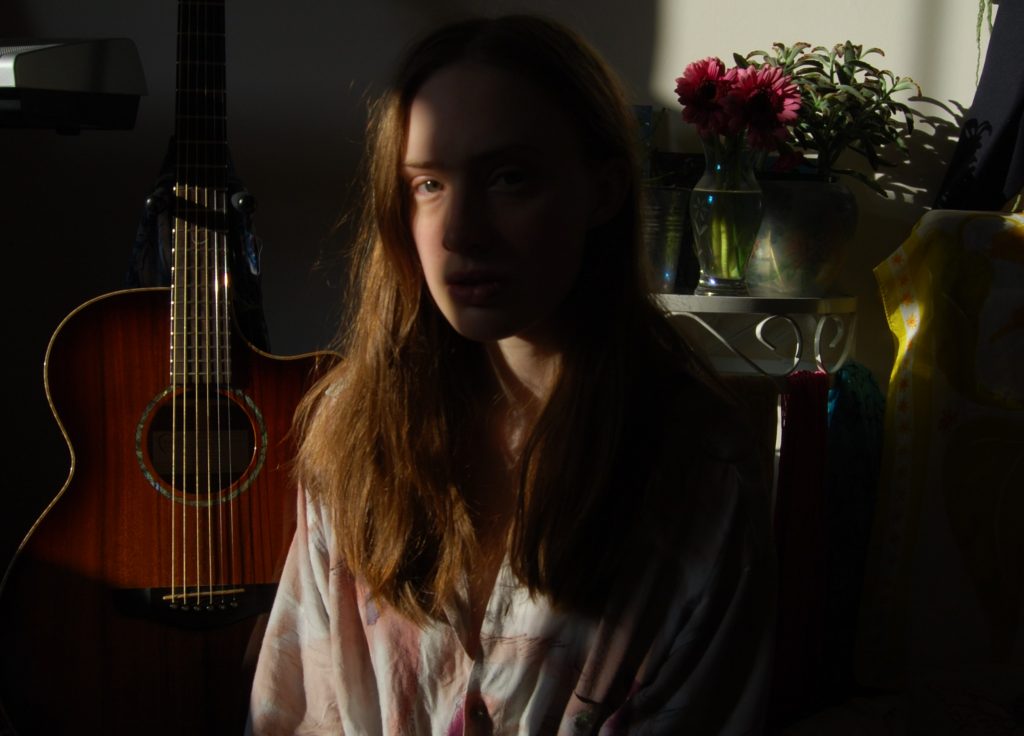 Photograph of Sakara in partial shade looking directly at the camera, the acoustic guitar and table of plants from previous photos are visible behind her.