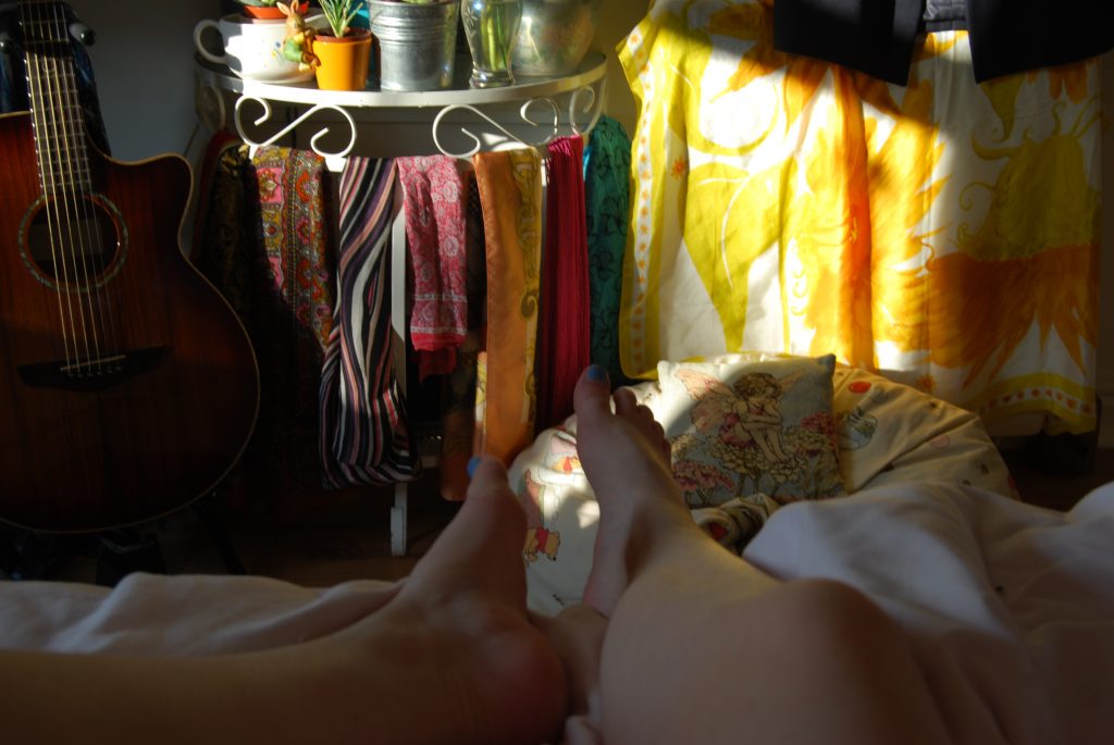 A photo lit by late afternoon sunlight slanting into the room. There is an acoustic guitar on the left hand side, a table next to it is covered in small plant pots and draped with brightly coloured scarves.