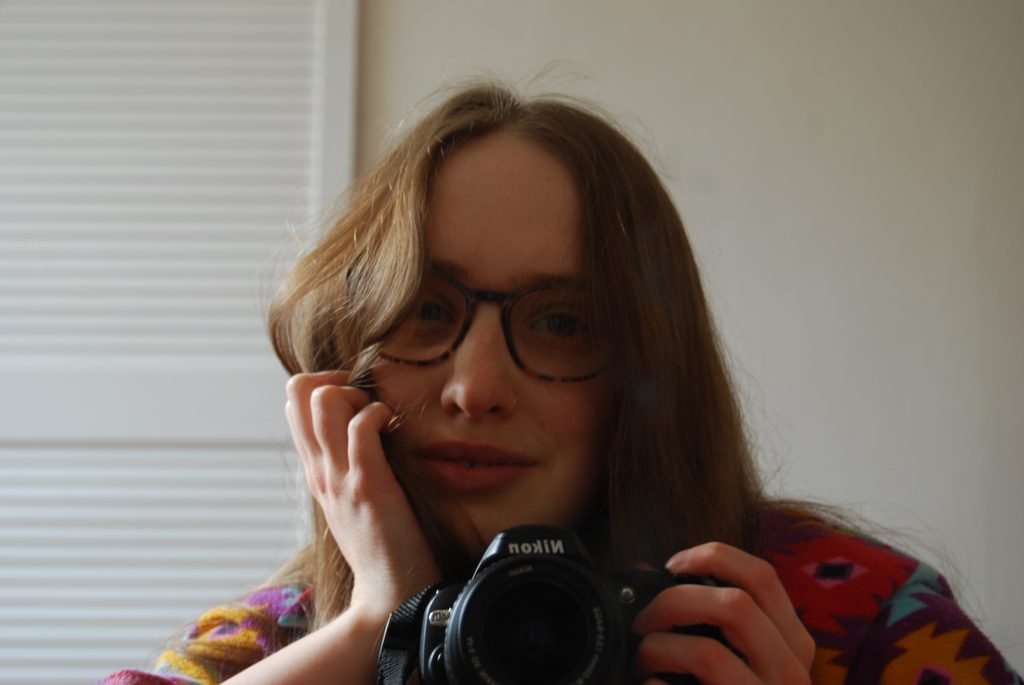 Mirror selfie of Sakara taken with a digital camera, she is smiling into the lens and is supporting her head with one hand up to her face, squishing some of her golden brown hair.