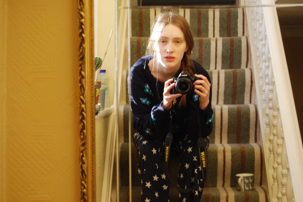 A mirror selfie of Sakara, a young white woman with light brown hair, sitting on the flight of stairs in her home. There is a striped carpet and a window to the left, white sunlight is coming through. It feels peaceful and a little hazy. There's a mug of tea on the step next to her.