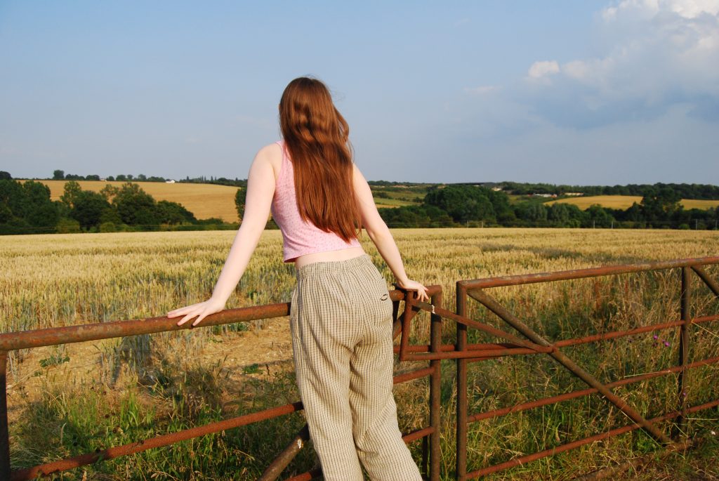 Sakara leaning forward on the gate with her back to the camera like she's balancing and looking out over the field.