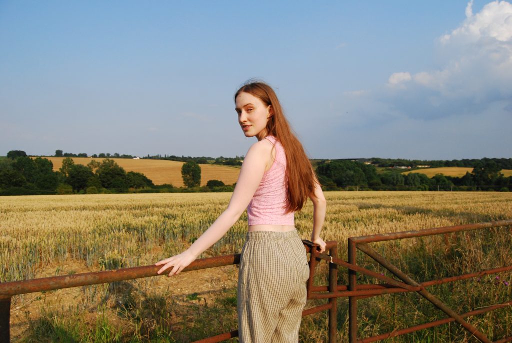 Sakara looks over her shoulder in front of the same field, she's wearing a tight pink crop top with little flowers printed on it and lose checked trousers with an elasticated waist.