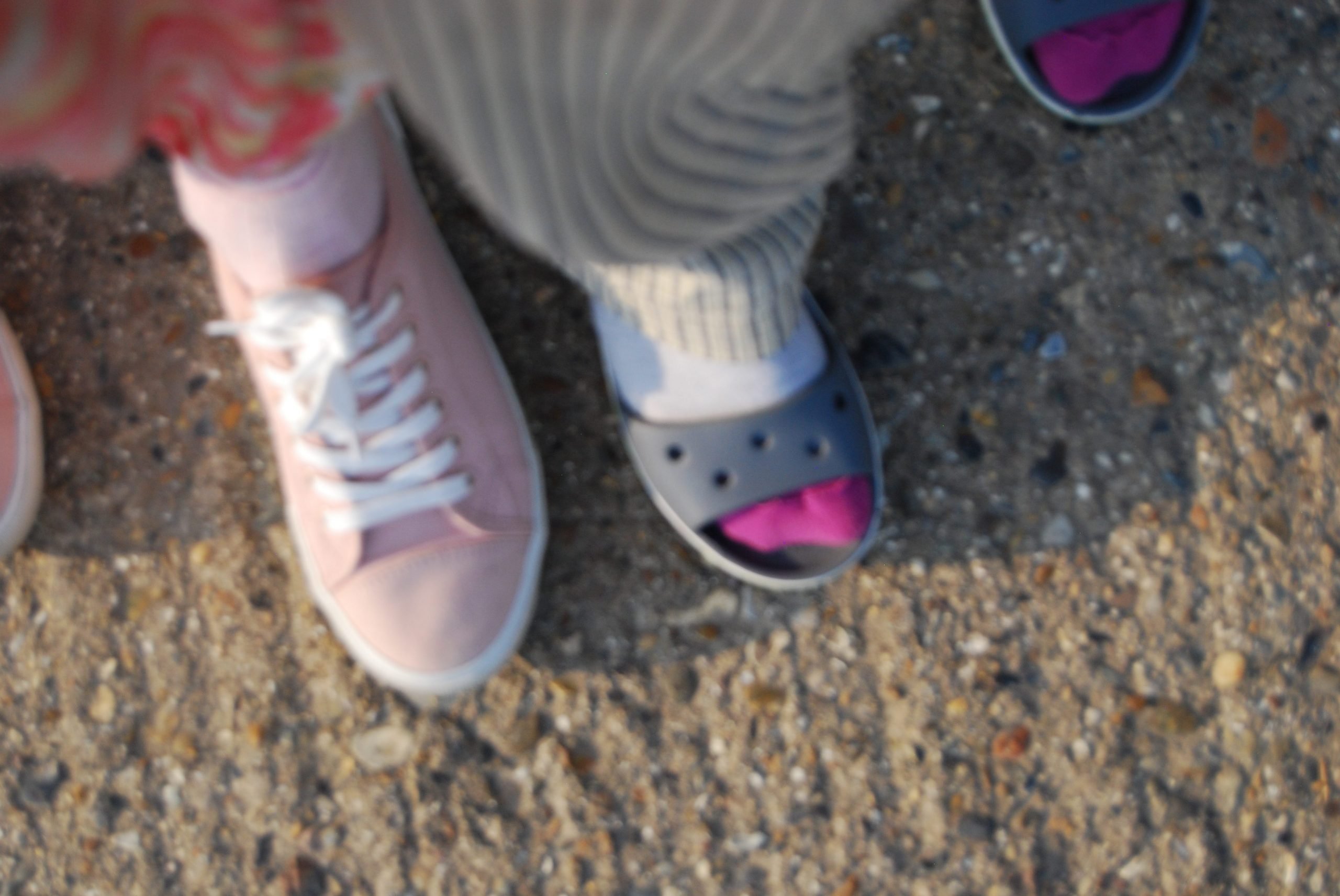 A blurry photo of two sets of feet side by side. One wears socks and sandals, the other converse-like pink shoes.