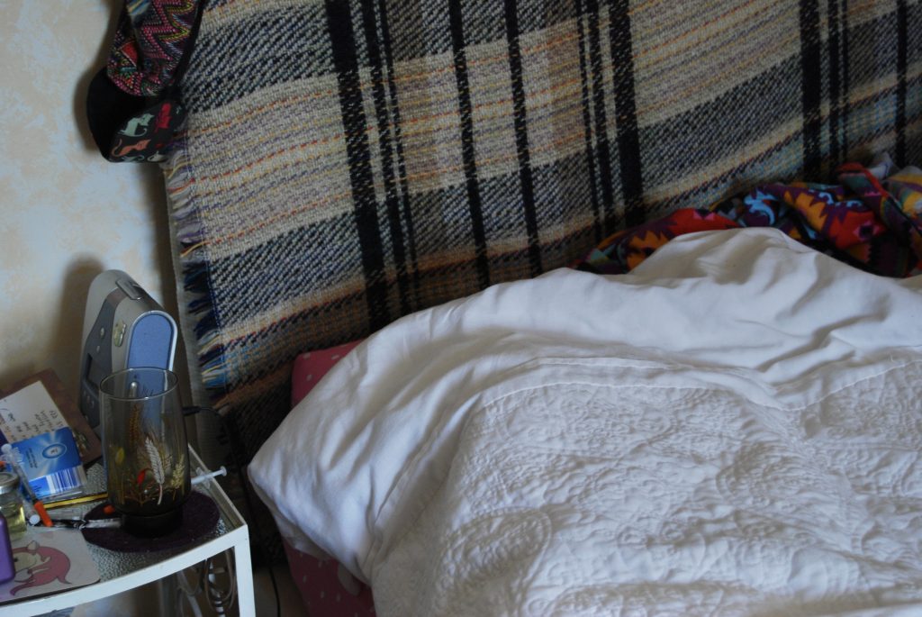 A close up image of the top of Sakara's bed, a headboard covered in a checked blanket is visible, along with a white quilt and screwed up colourful shirt. Her bedside table has medication and a wave noise player on it.