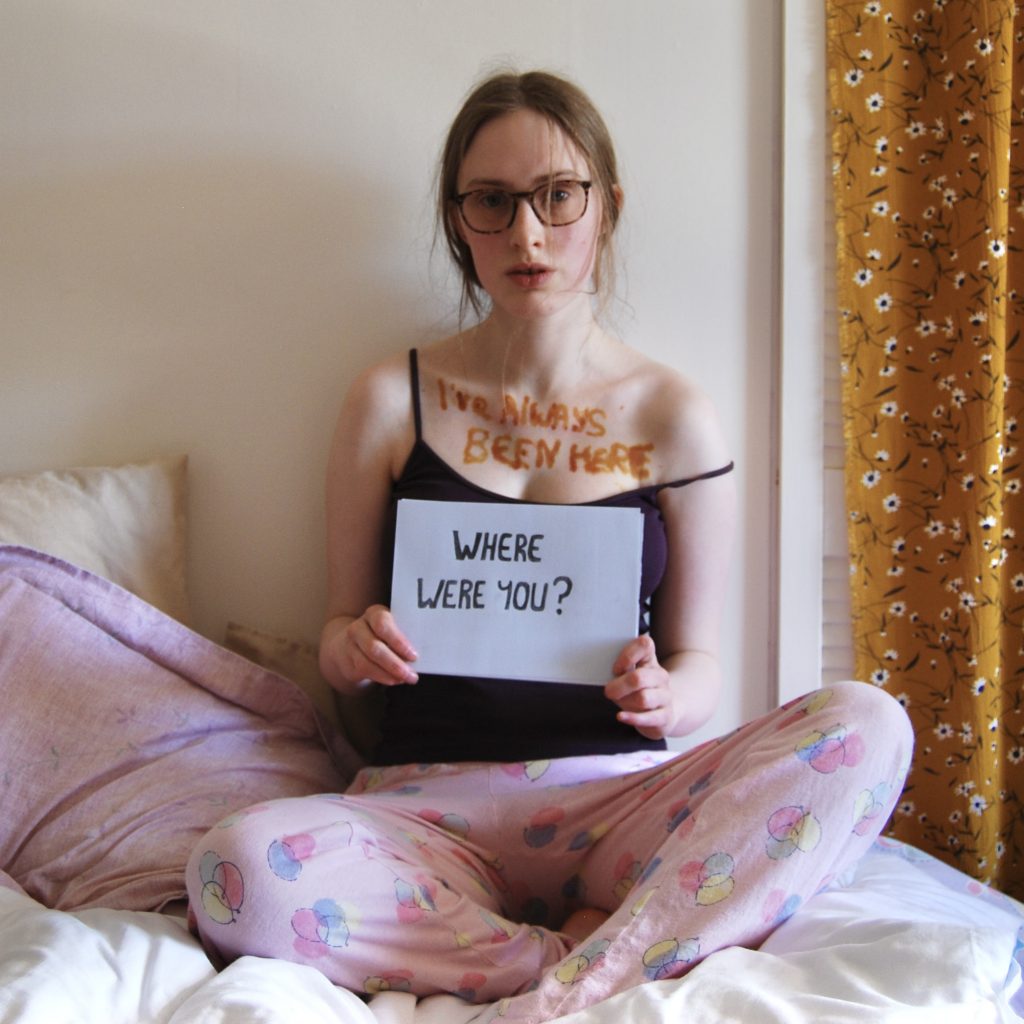 Sakara sits on her bed with the "where were you?" sign in her hands.