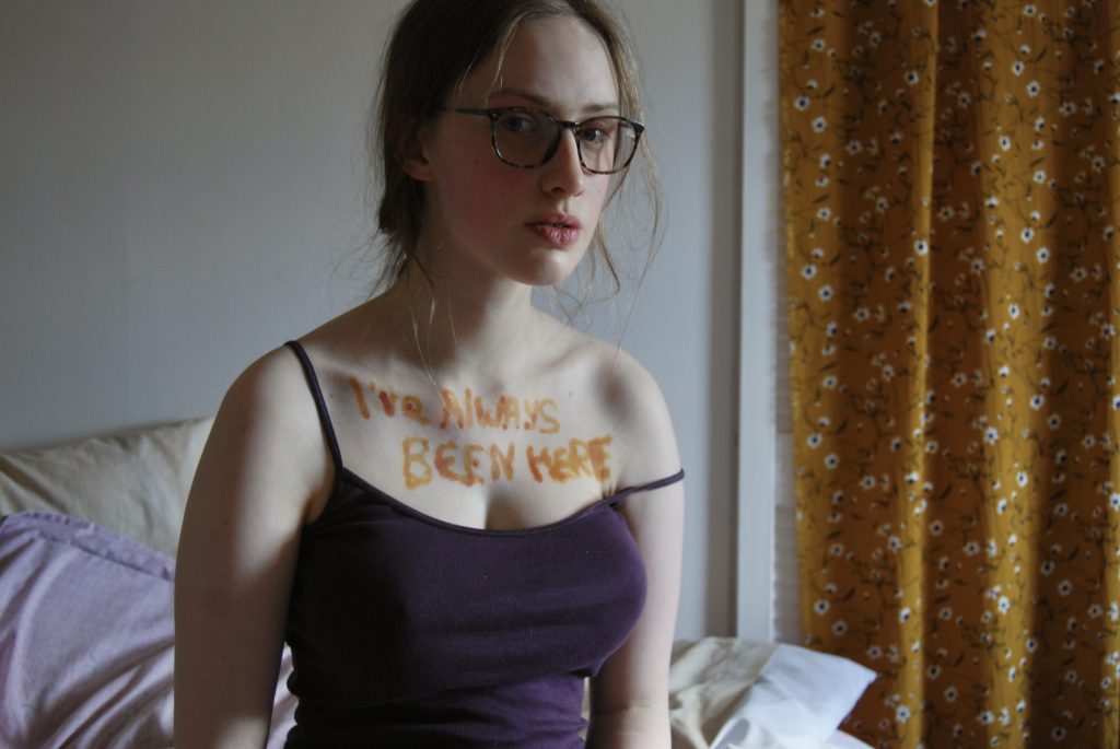 One final photo of Sakara looking into the camera, not holding a sign but still with "I've always been here" written across her chest.
