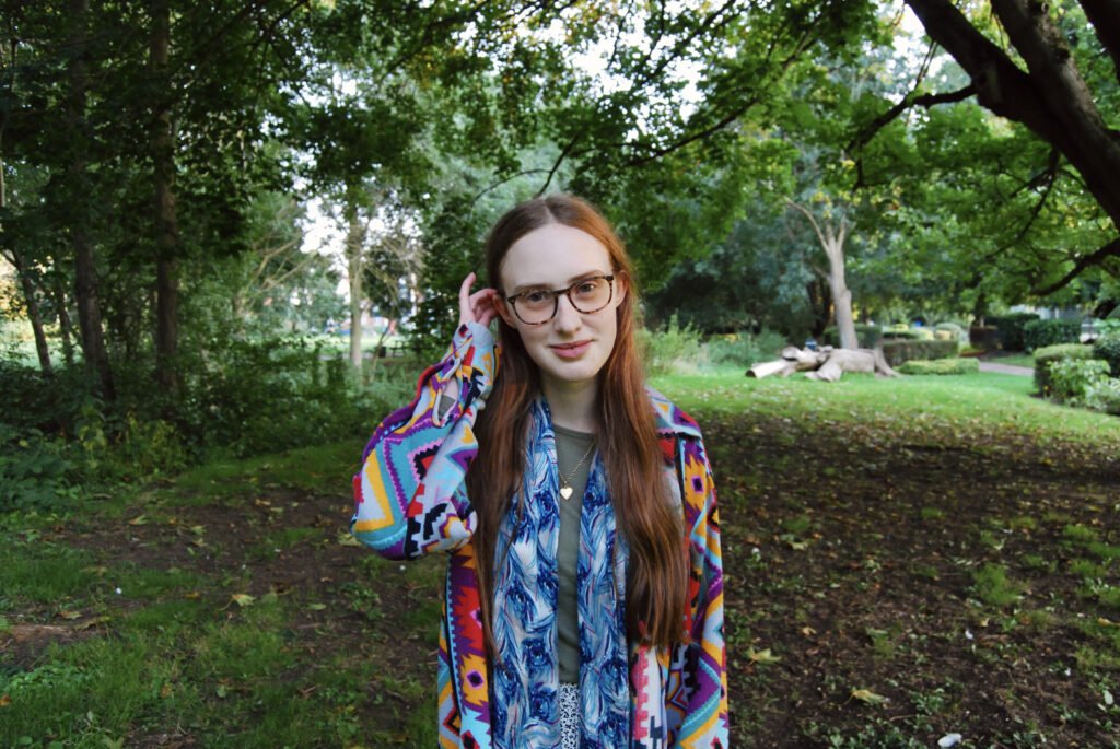 Photo of Sakara standing near trees, she is pushing her hair behind her ear and wears a colourful top.