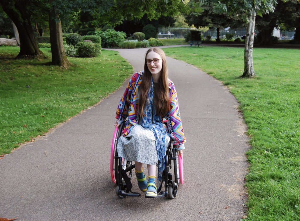 Another photo of Sakara in her wheelchair. She looks happy and is leaning to one side, pushing one wheel forwards.