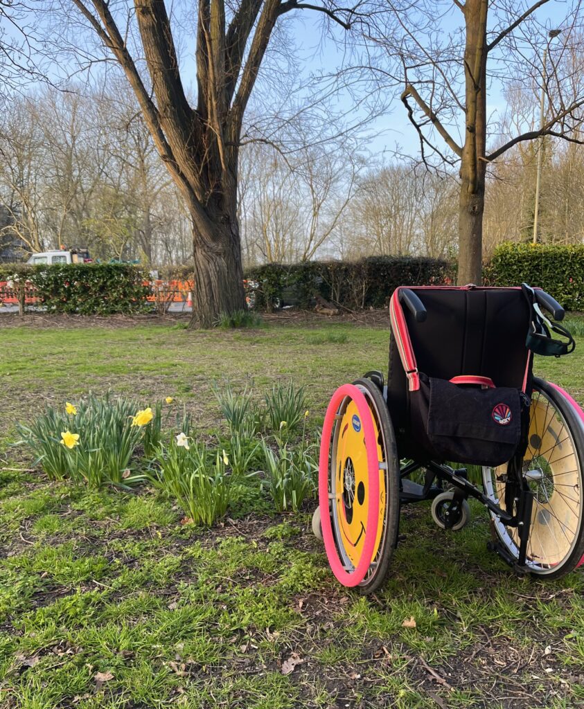 The wheelchair shown from behind. It has yellow smiley face wheel covers and pink wheel rims, a bag is hung over the handles.
